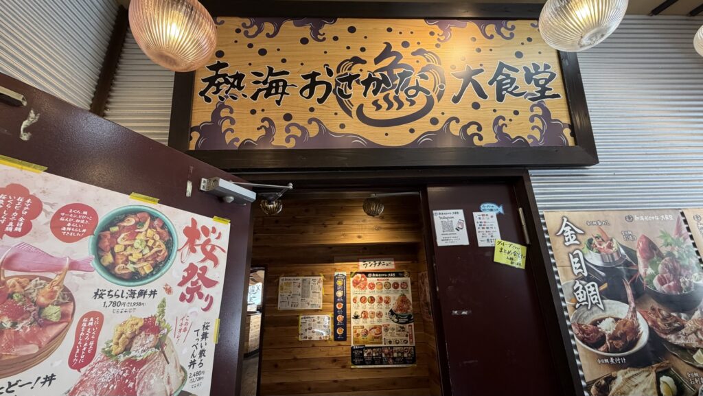 「熱海おさかな大食堂」の入り口上部に掲げられた、ダイナミックな波のイラスト入り木製看板。ドアには「桜祭り」や「金目鯛」の大きなポスターが貼られ、活気ある外観
