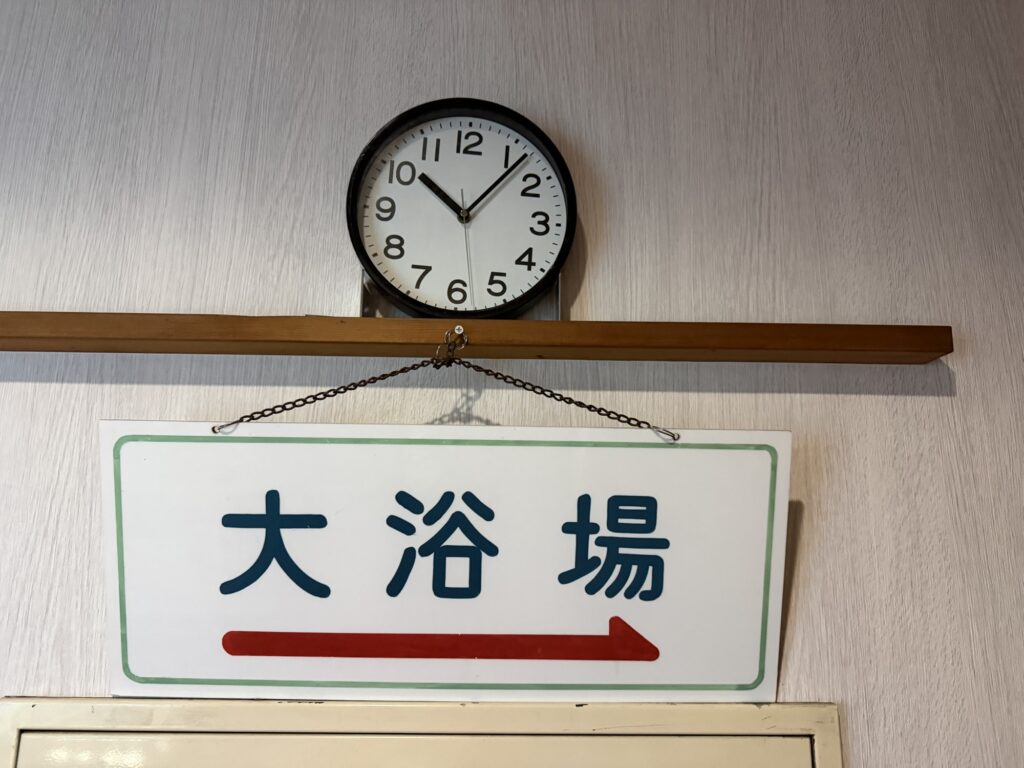 湯ーとぴあ館内の「大浴場」と矢印が書かれた案内板と壁時計