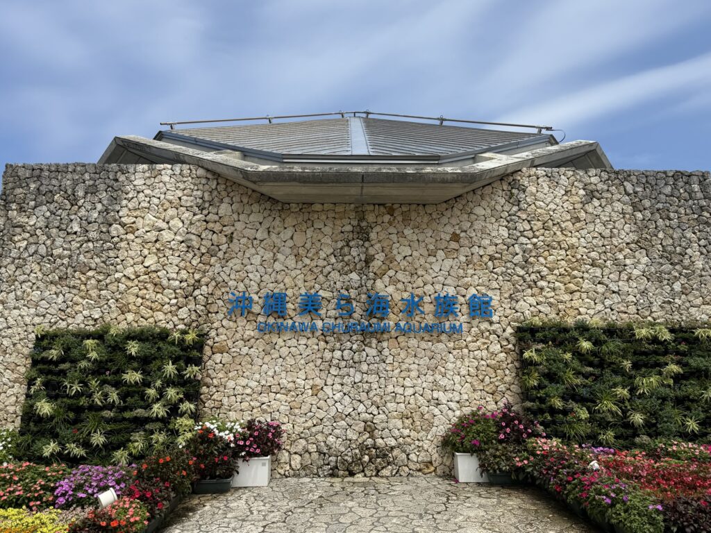 石積みの壁に「沖縄美ら海水族館」の文字が掲げられた建物入口、花壇に囲まれたフォトスポット